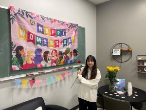 International student holding a rose and standing in front of the Women's Day banner in the Office of International Programs.