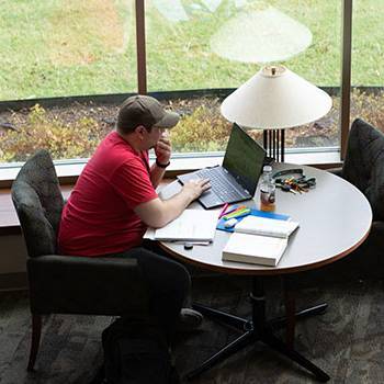 Student studying at table with books and computer on BA campus.